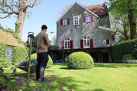 Eine Person steht in einem Garten mit Formschnittgehölzen und Bäumen. Sie blickt auf das im Kern mittelalterliche Gebäude. Die Person steuert mit einer Fernbedienung eine Drohne, die direkt vor ihr in der Luft schwebt.